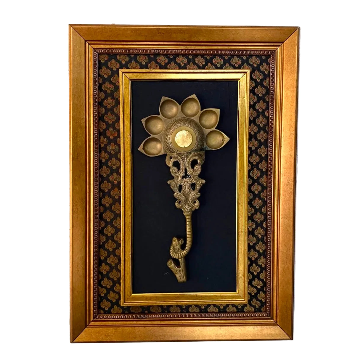 A framed vintage brass wall hook with a lotus flower head and elephant trunk, mounted on a black patterned background inside a gold frame, hung on a white textured wall.
