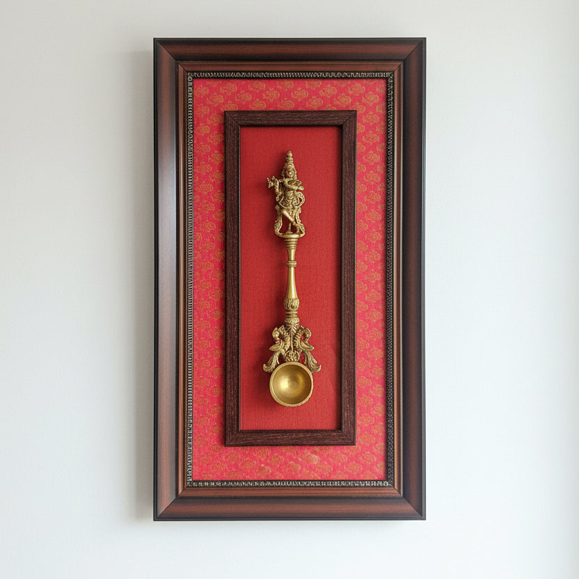 A framed decorative object featuring a gold-toned ornate spoon-like sculpture mounted on a red patterned background, enclosed in a dark wood frame. The frame is being held against a real indoor background with shelves and papers visible.