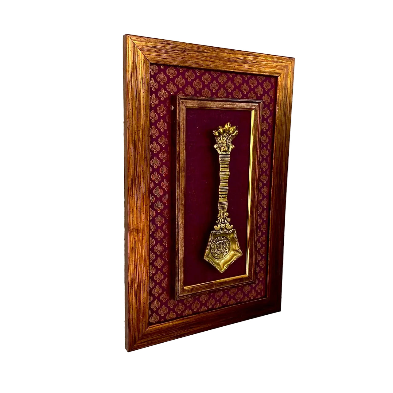 A framed decorative wall piece featuring a gold-colored miniature object mounted on a deep burgundy patterned fabric background, enclosed in a medium brown wooden frame and hung on a textured white wall.