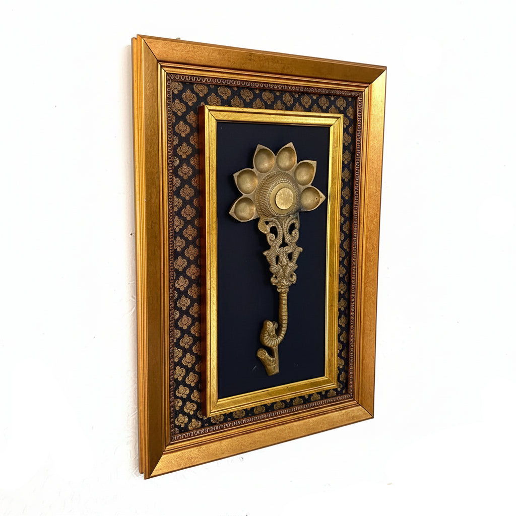 A framed decorative wall piece featuring an ornate gold-toned flower-shaped wall hook mounted on a black patterned background, enclosed in a gold frame and hung on a textured white wall.