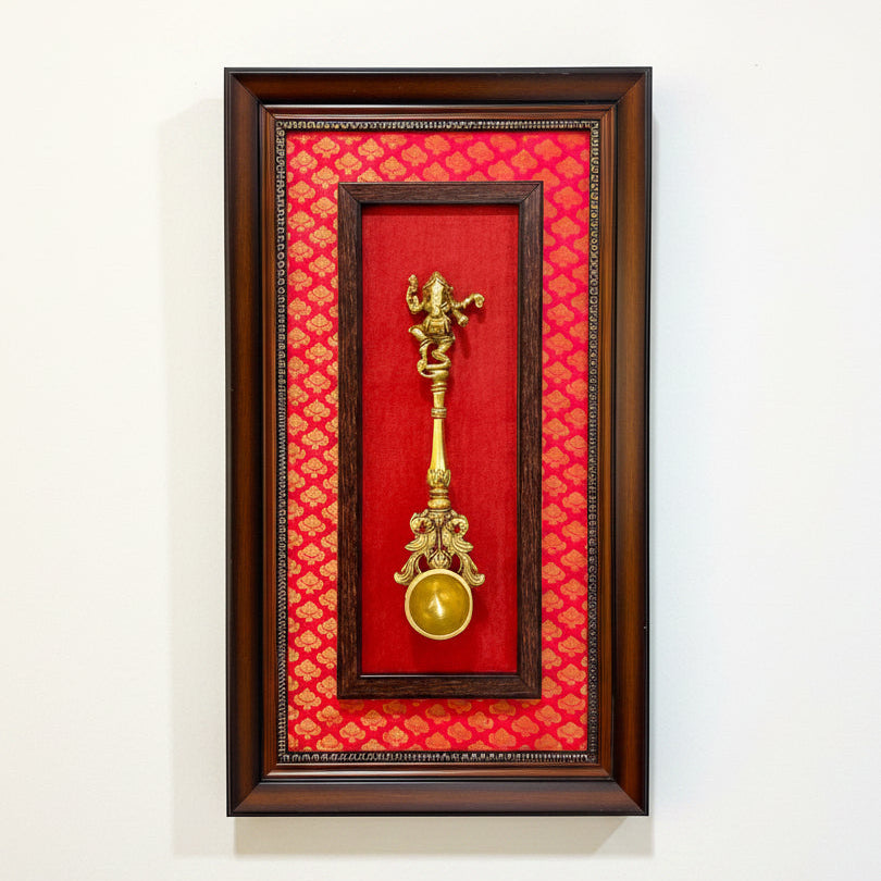 A framed wall art piece featuring a brass Ganesha lamp (Lantana) mounted on a vibrant red patterned fabric background, enclosed in a dark brown wooden frame and hung on a textured off-white wall.