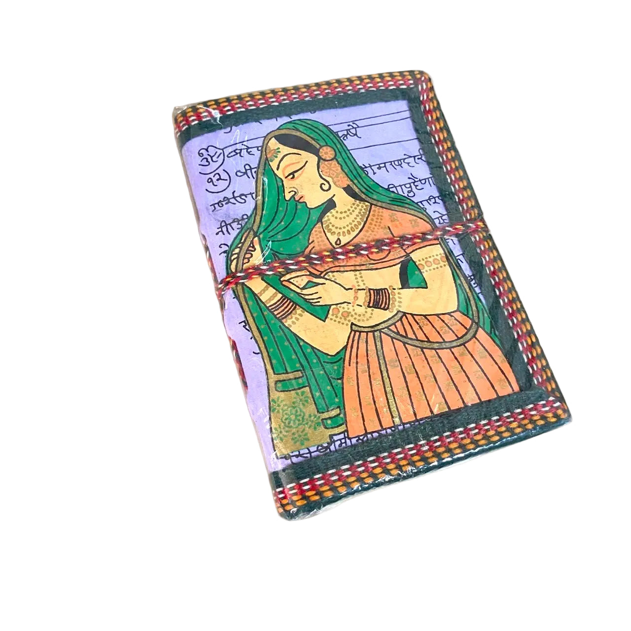 A small rectangular notebook or journal with a hand-painted traditional Indian style illustration of a woman in green and orange clothing, bound with a red and gold patterned string, resting on a light wood surface.