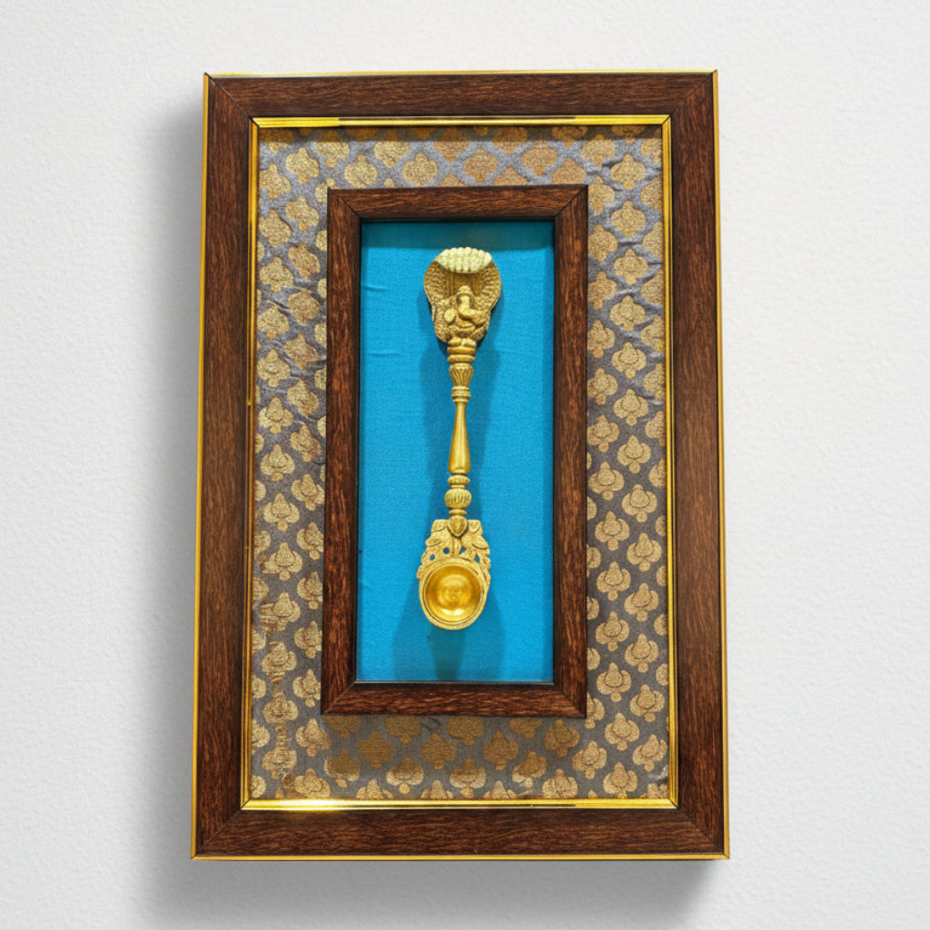 A framed decorative object featuring a gold-toned ornate spoon or ladle mounted against a bright blue backing, surrounded by patterned blue-and-gold fabric and a dark wood frame with gold trim, shown on a white background.