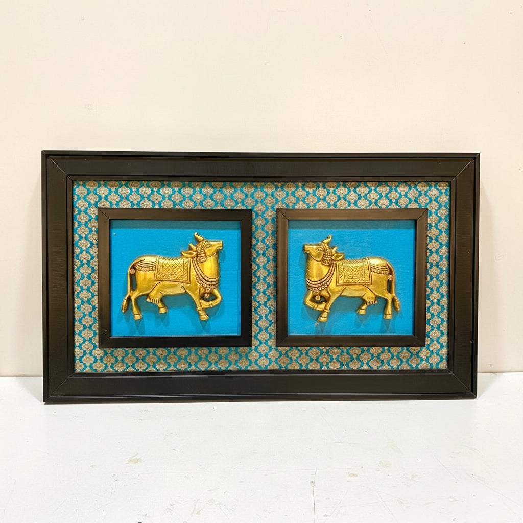 A framed wall art piece featuring two gold-colored bull sculptures on a bright turquoise background with a teal and gold patterned border, enclosed in a dark wood frame.