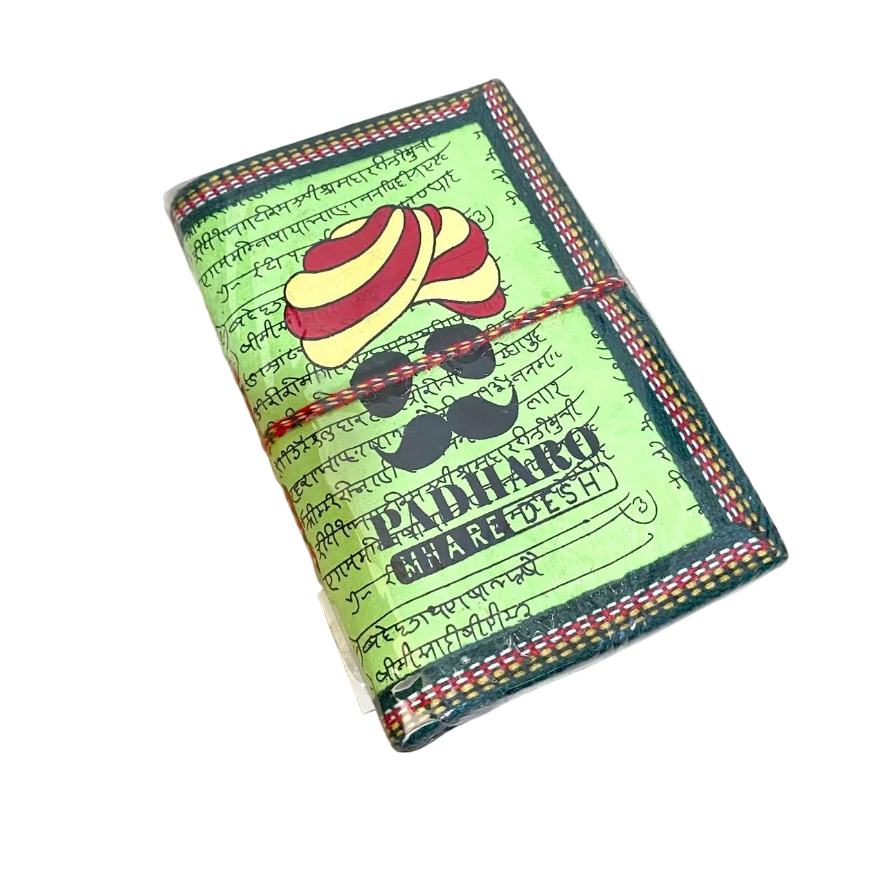 A handmade paper wallet or notebook cover featuring a green background with black script, a red and yellow striped turban illustration, black mustache graphic, and the text 'PADIHARO CHAREDES' in English and Devanagari script. The item is wrapped with a red and black string and photographed on a light wood tabletop.