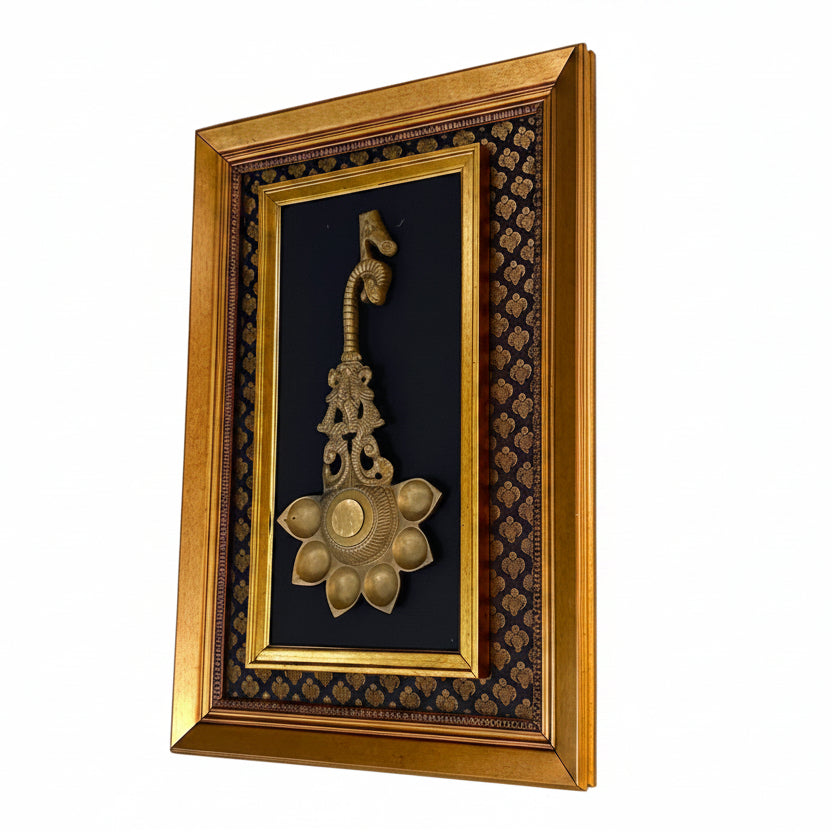 A framed decorative wall piece featuring an ornate gold-toned metal ornament mounted on a black patterned background, enclosed in a wide gold frame and hung on a textured white wall.