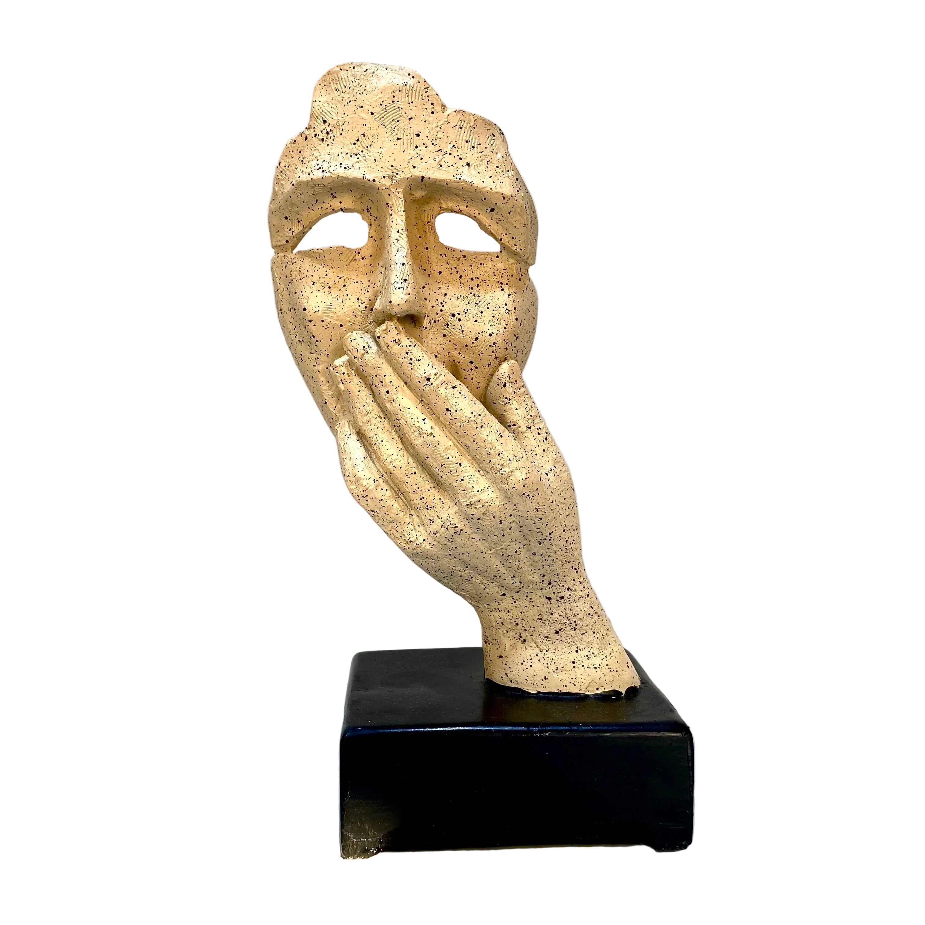 Decorative sculpture of a hand covering a mask on a black base against a textured wall.