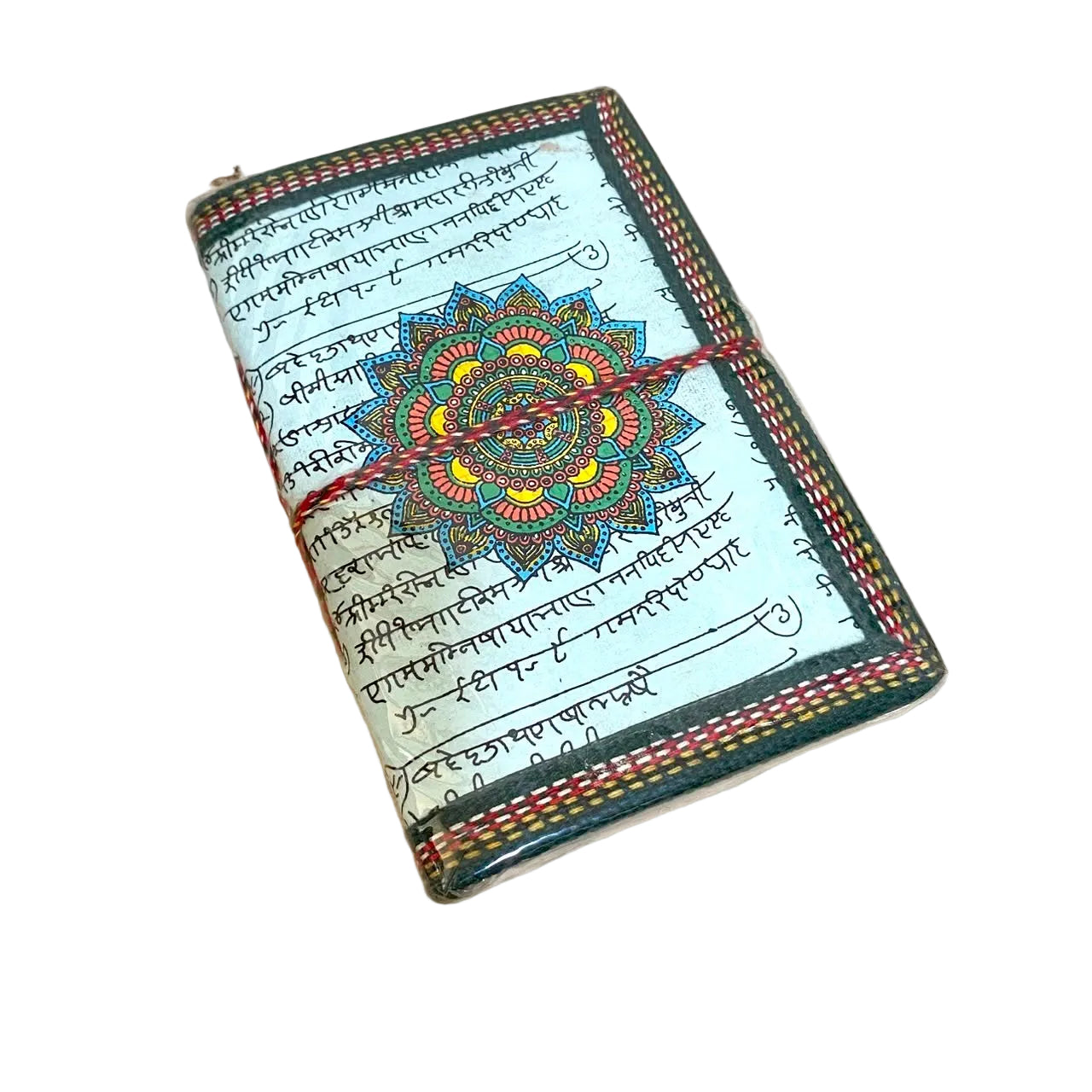 A small rectangular notebook or journal lying on a light wood tabletop. The cover has a pale blue background with black script, a large colorful mandala design in the center, and a dark green stitched border with red and yellow accents.
