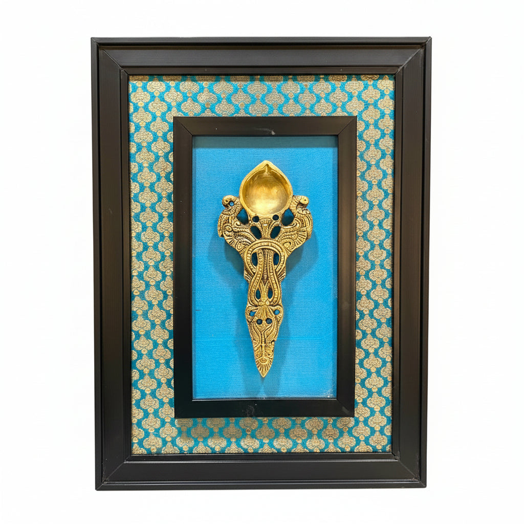 A framed decorative wall piece featuring a gold-toned ornate spoon mounted on a bright blue patterned fabric background, enclosed in a black frame.