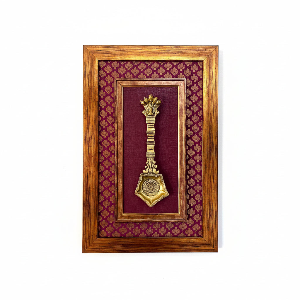 A framed decorative wall piece featuring a gold-toned ornamental key or latch mounted on a deep burgundy fabric background with a repeating gold pattern, enclosed in a medium brown wooden frame and hung on a white textured wall.