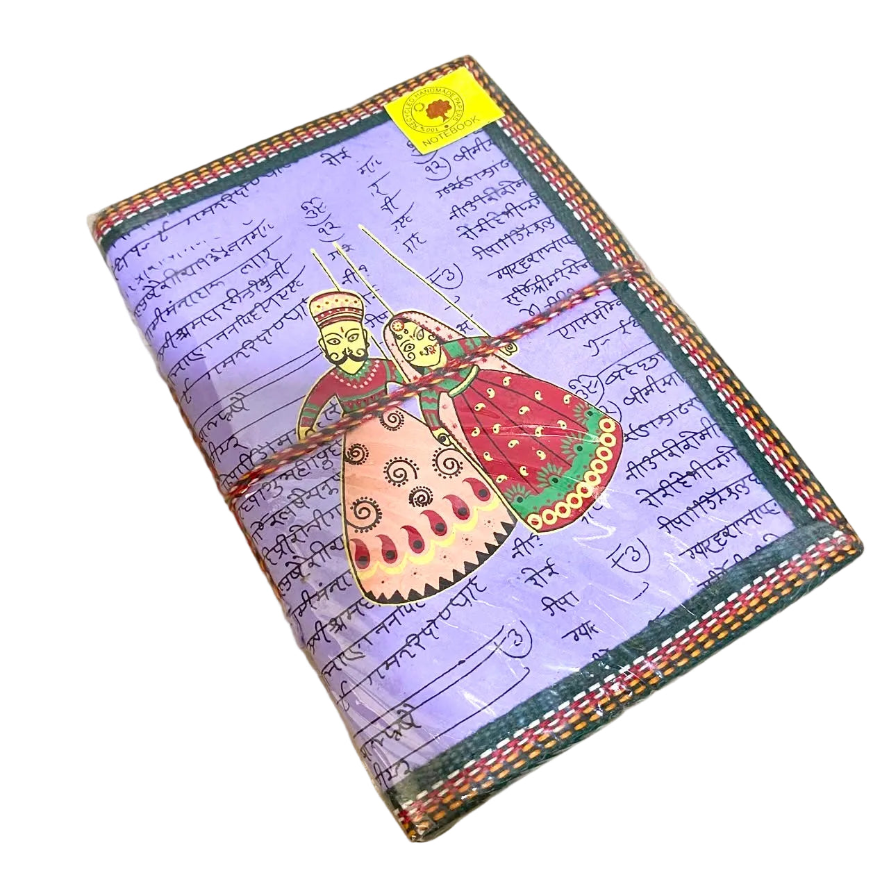 A square decorative notebook or journal lying on a light wood surface. The cover has a lavender background with handwritten script, a colorful illustration of two figures in traditional clothing, and a multicolored stitched border.