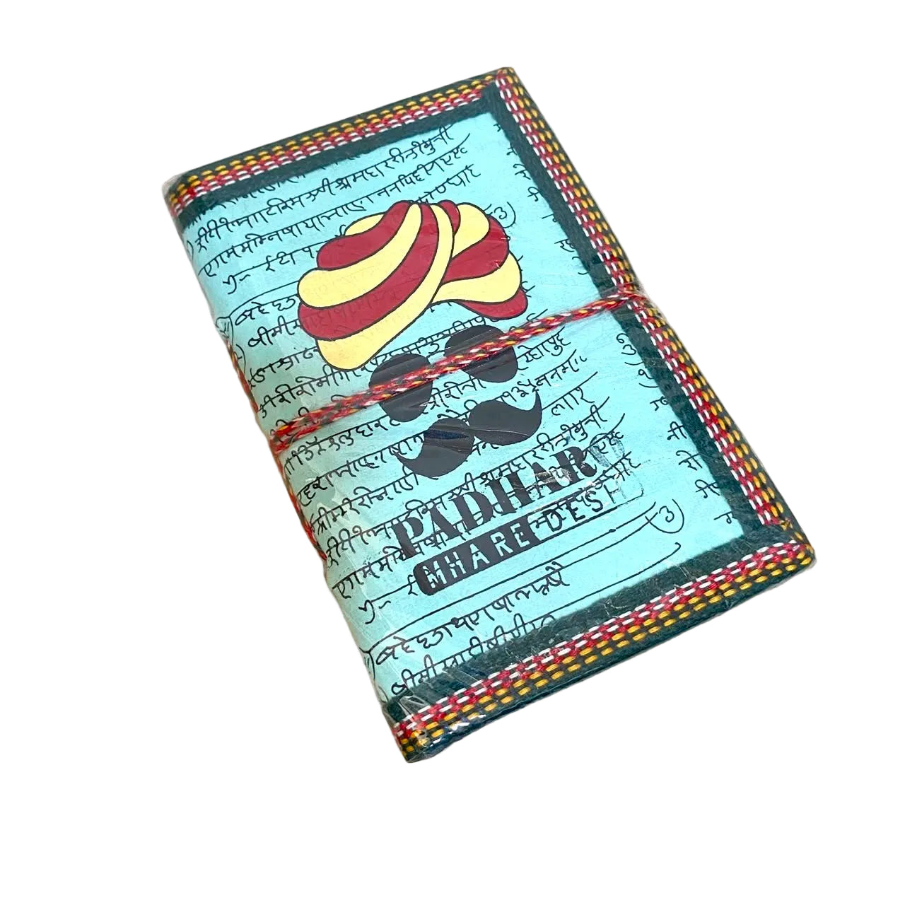 A handmade wallet or notebook cover featuring a light blue background with black script, a yellow and red striped turban illustration, a black mustache graphic, and the text 'PUDHAM KHAREDES'. It has a multicolor braided cord tied around it and is placed on a light wood surface.