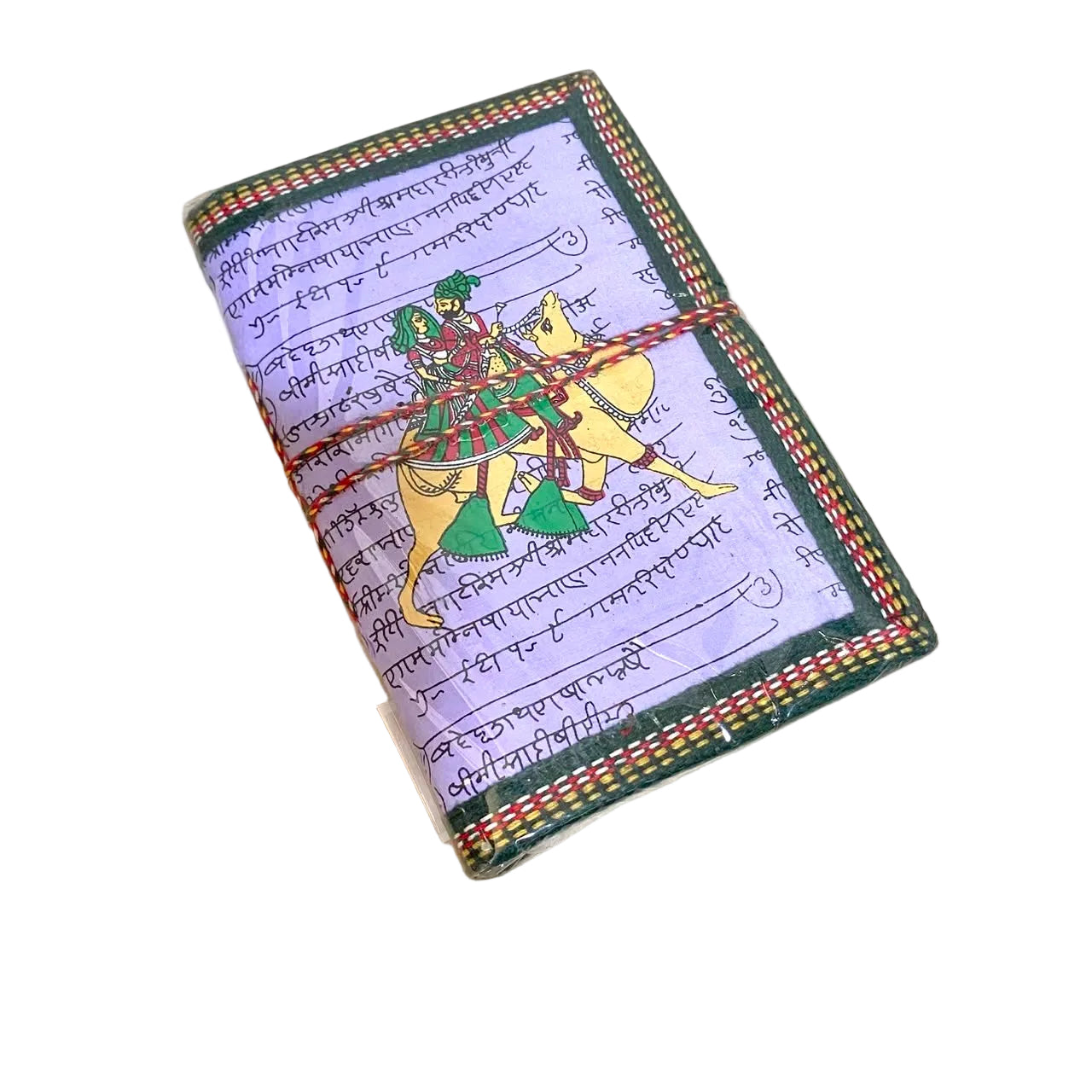 A small rectangular notebook or journal lying on a light wood tabletop. The cover has a pale lavender background with printed script, a dark green border, and a colorful illustration of a rider on a yellow horse.